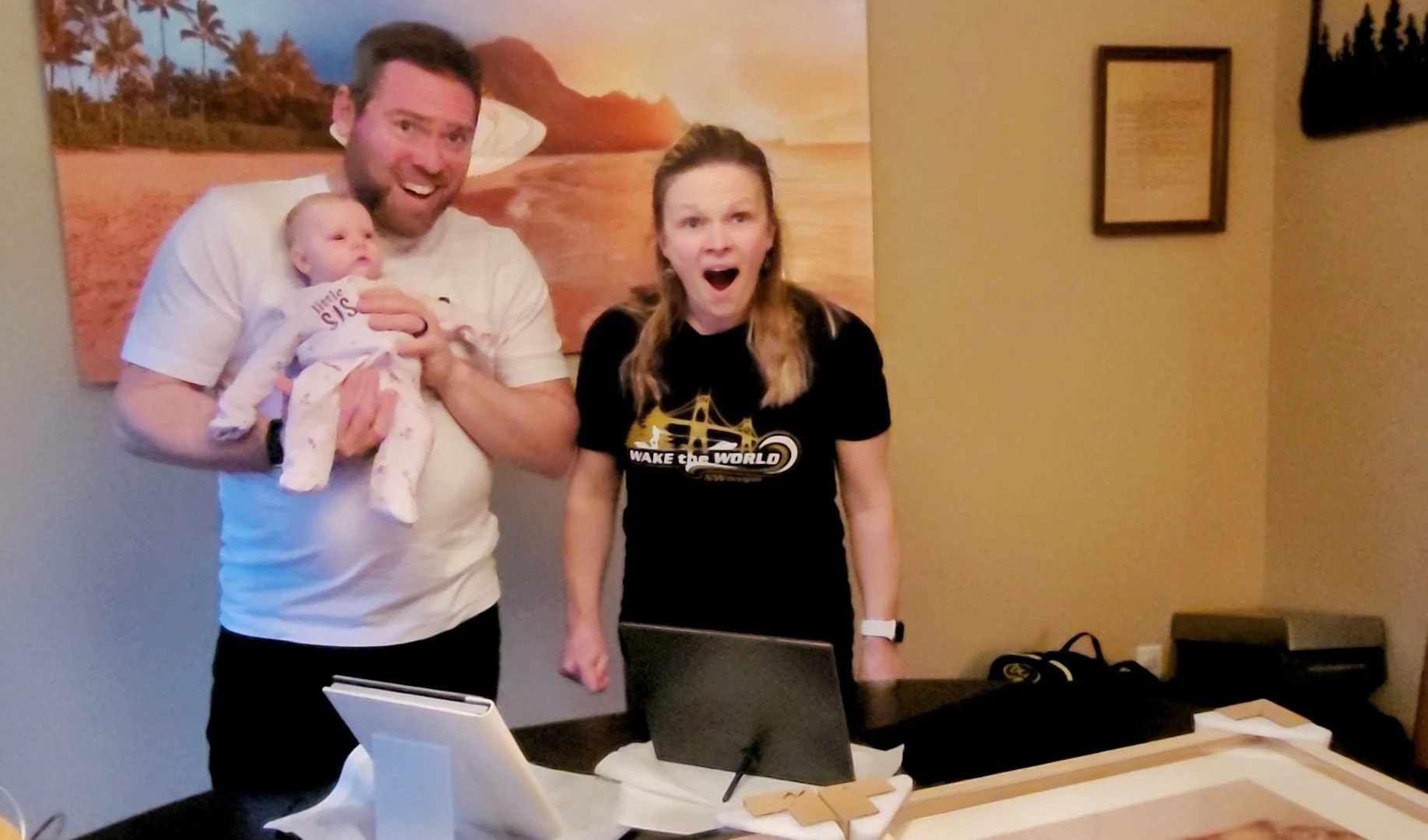 A candid shot of a surprised and happy couple holding their baby during a photo gallery reveal session. The mother has an excited expression while the father holds their infant, showcasing the emotional experience of viewing final portraits with a Po