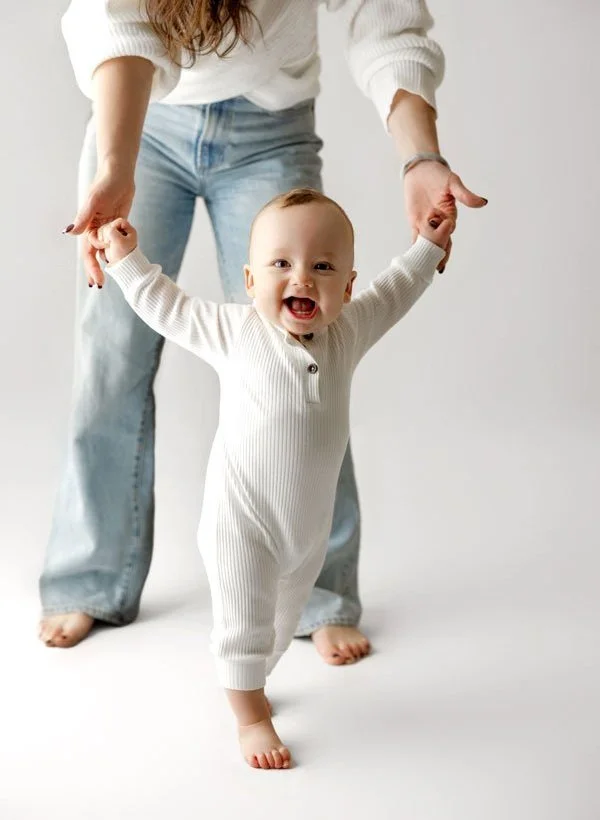 A happy, smiling baby in a white ribbed sleeper holding an adult's hands while taking first steps in a bright studio with a high-key white background.