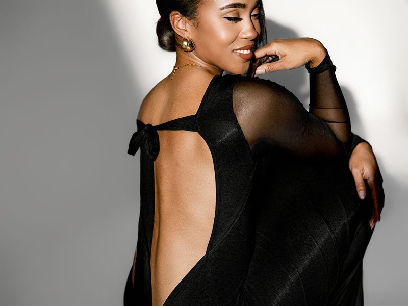 A close-up studio portrait of a woman in an elegant black backless dress, looking over her shoulder with a soft smile against a minimalist grey background with dramatic lighting.