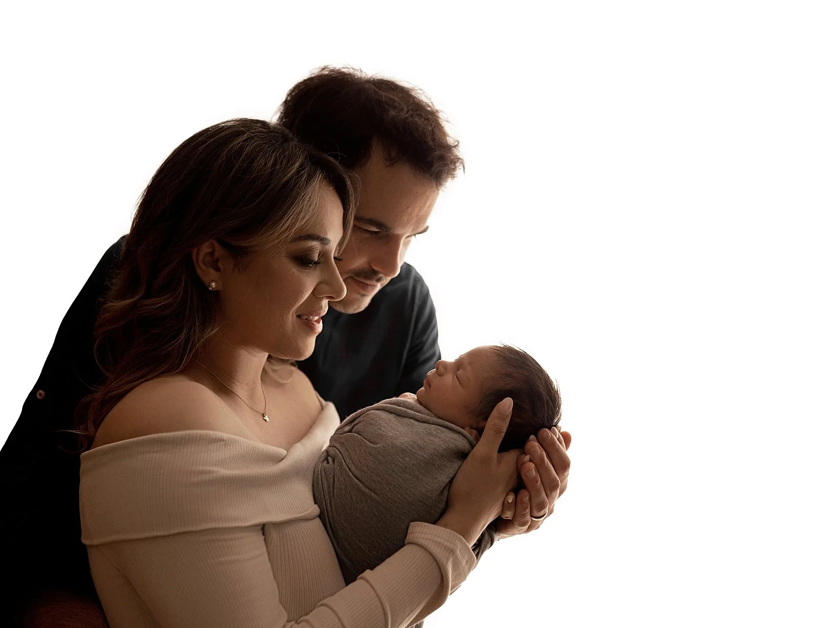 A professional studio newborn portrait featuring a mother and father looking down lovingly at their sleeping infant. The baby is snugly wrapped in a neutral gray swaddle and held gently in the mother's hands. The image uses a dramatic backlit lightin