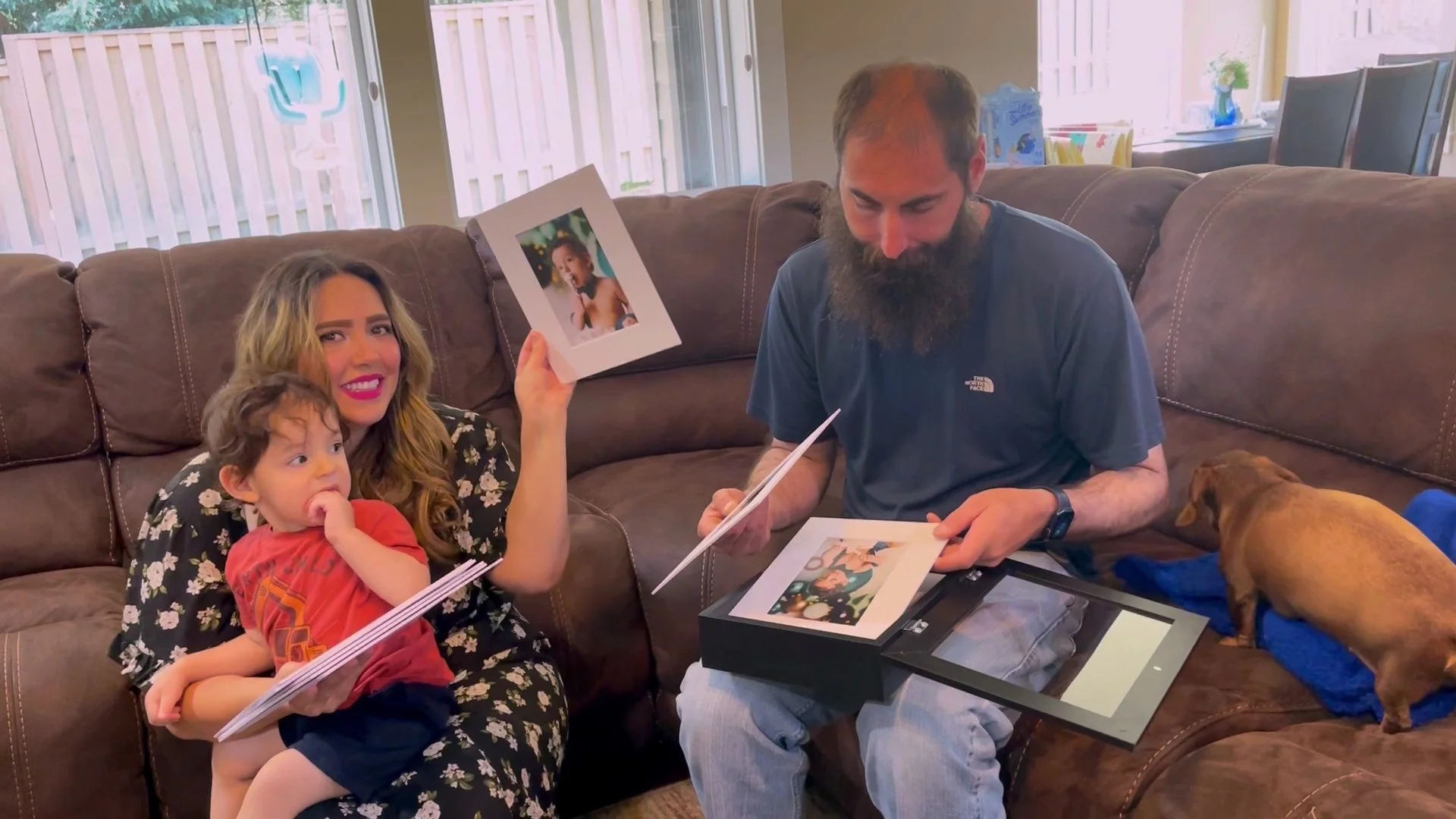 A happy family—mother, father, and young son—sits together on a couch viewing high-quality matted photo prints from a professional reveal session. The mother holds up a matted portrait while the father looks through a presentation box, showcasing the