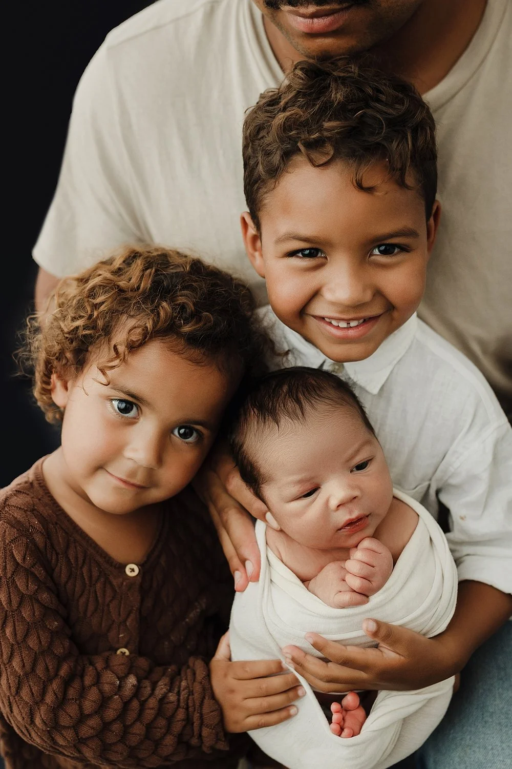 A heartwarming sibling photograph featuring a young boy with curly hair in a white button-down shirt and a younger girl with curly brown hair in a brown knit cardigan. Together, they are carefully holding a newborn baby who is snugly swaddled in a wh