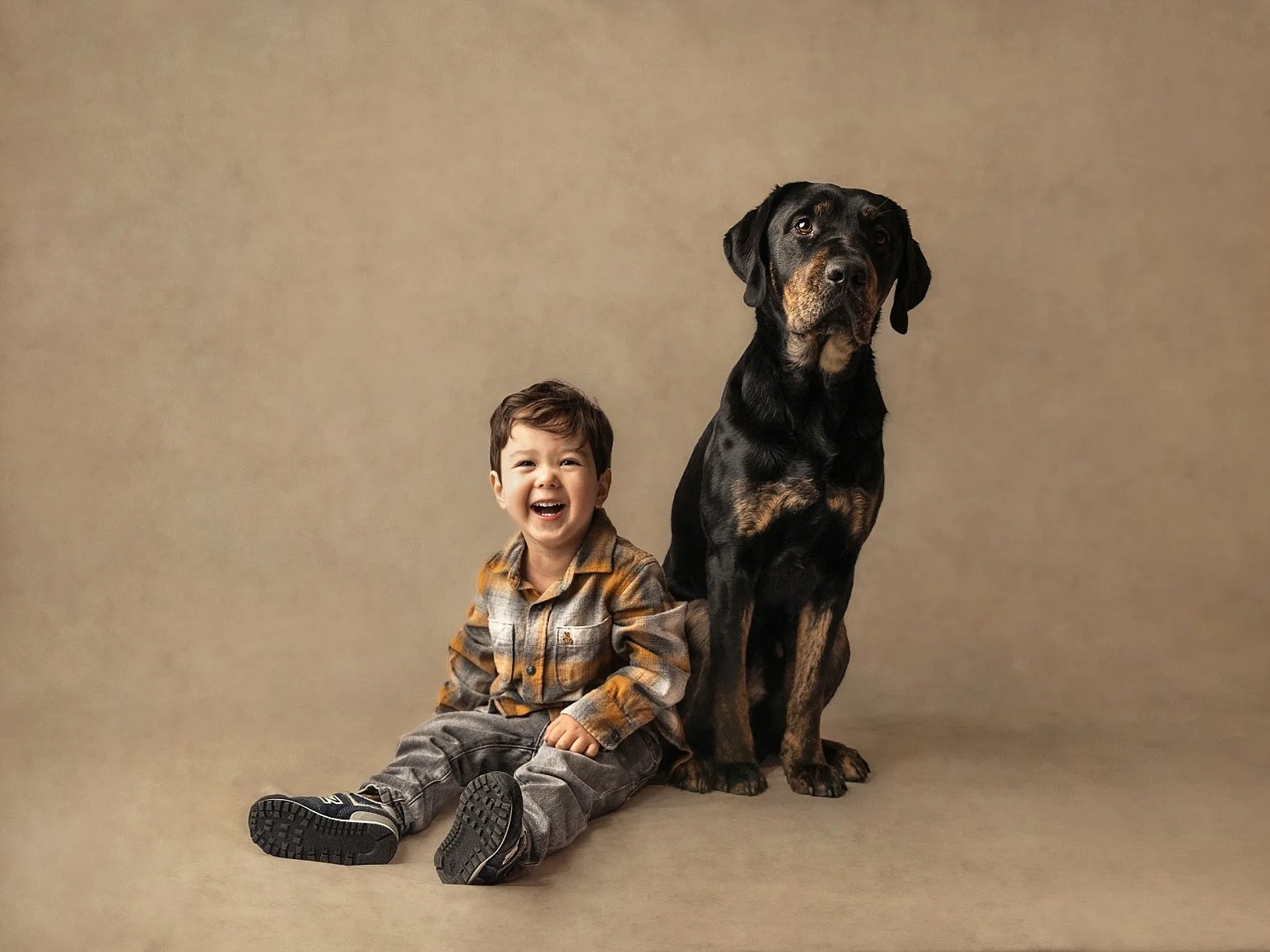 This soul-stirring fine art portrait features a candid moment of joy between a toddler and his large canine companion. The boy is captured laughing heartily, dressed in a classic flannel shirt and denim, while the dog sits tall and protective by his 