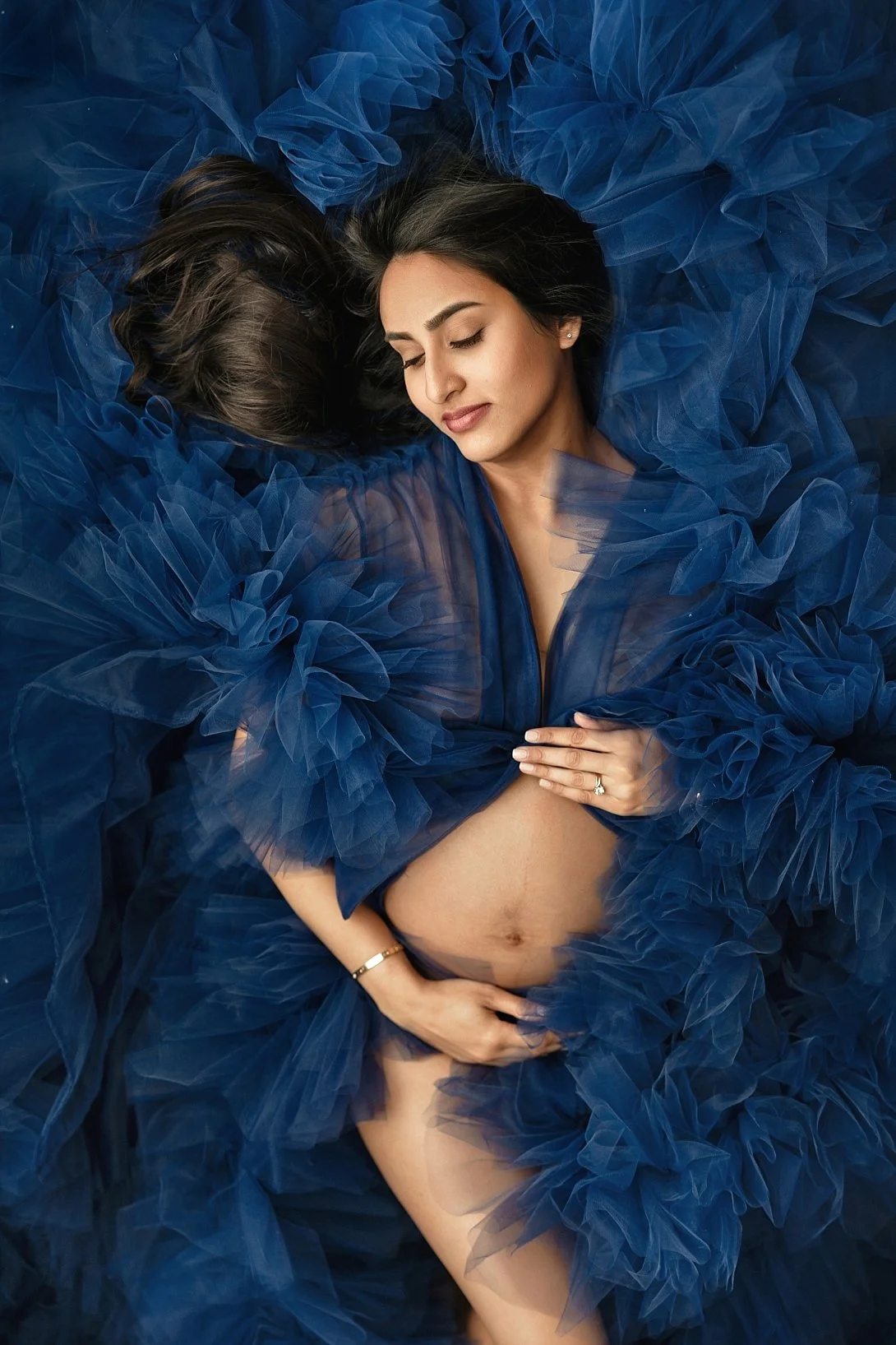 Elegant maternity portrait in Portland, Oregon, featuring a pregnant woman lying amongst a voluminous, ruffled blue tulle gown, gently holding her baby bump from a top-down view.