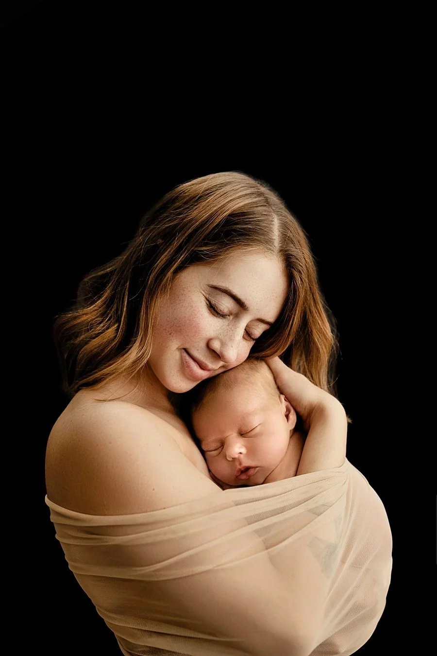 Fine-art studio portrait of a mother gently cradling her sleeping newborn baby wrapped in sheer fabric against a black backdrop in West Linn, Oregon.