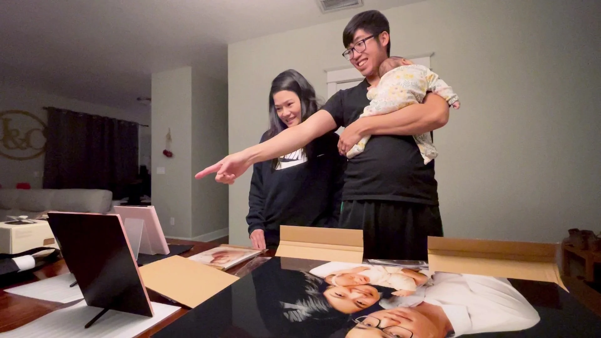 A young couple smiles as they view their new custom-framed family portraits during a gallery reveal session. The father holds a sleeping baby and points happily at the high-quality prints, showcasing the personalized service of a Portland family phot
