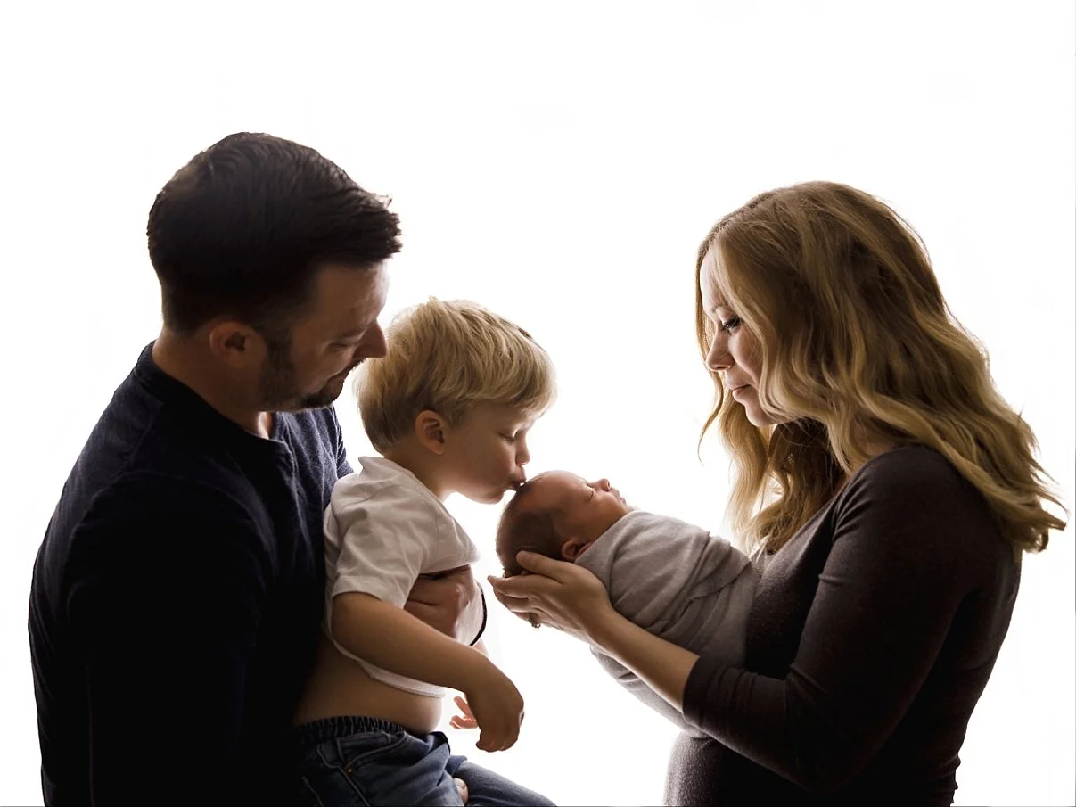 A professional high-contrast family photograph featuring a mother on the right in a dark long-sleeve top, cradling a newborn swaddled in grey. An older blonde toddler, held by his father on the left, leans in to gently kiss the infant's forehead. The
