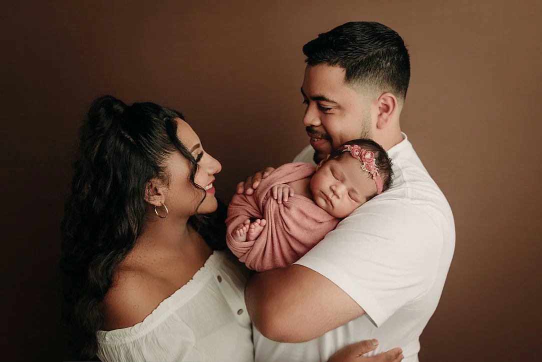 A tender family photograph featuring a mother with long dark curly hair and a father with a short beard looking at one another with affection. The father holds their infant daughter, who is peacefully asleep and swaddled in a dusty rose wrap with a m