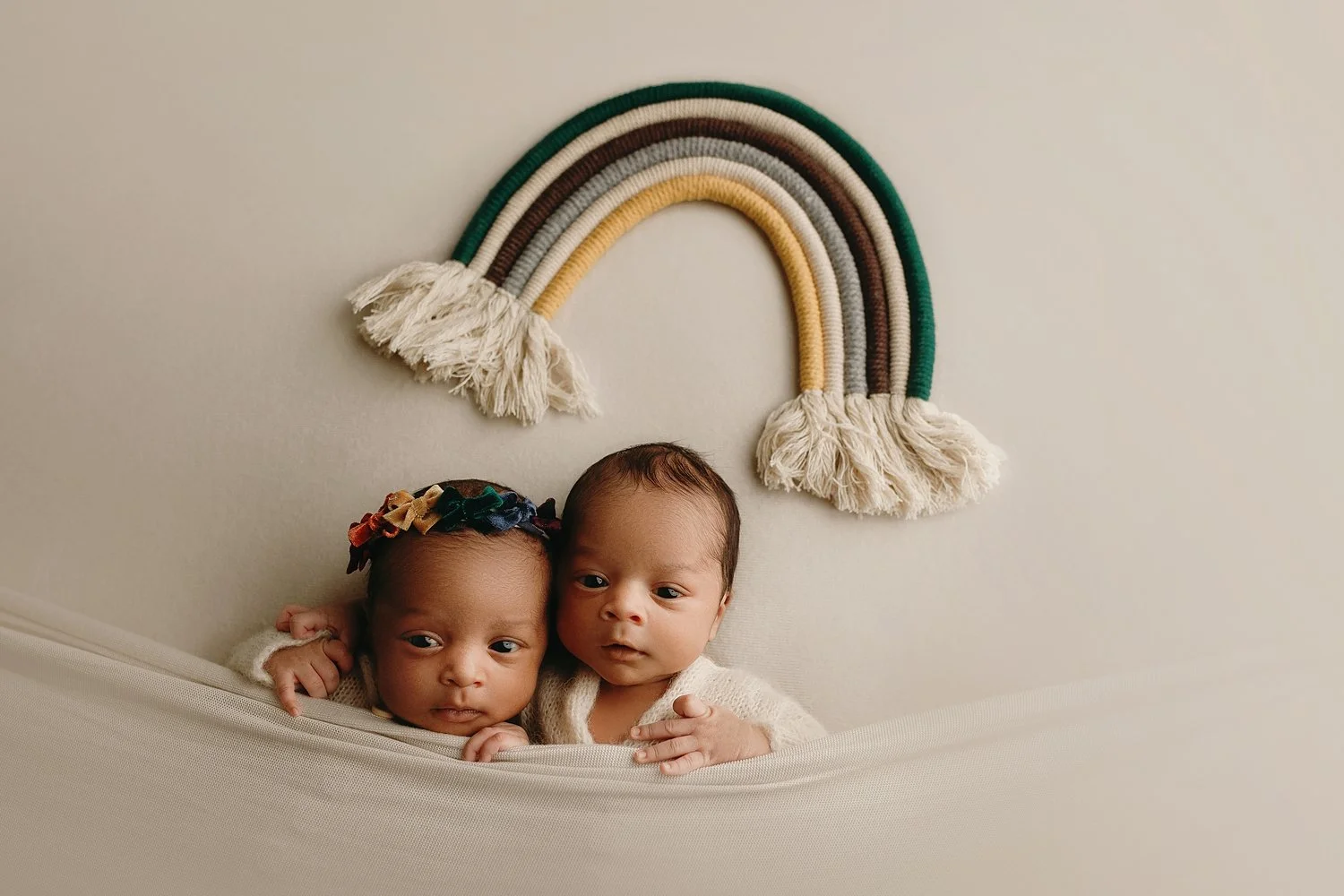 Newborn twins of NBA star Damian Lillard posing together in a cream-colored hammock during a professional photoshoot by Sunnymelon Photography. The scene features a boho-style macramé rainbow wall hanging and earthy, neutral tones.