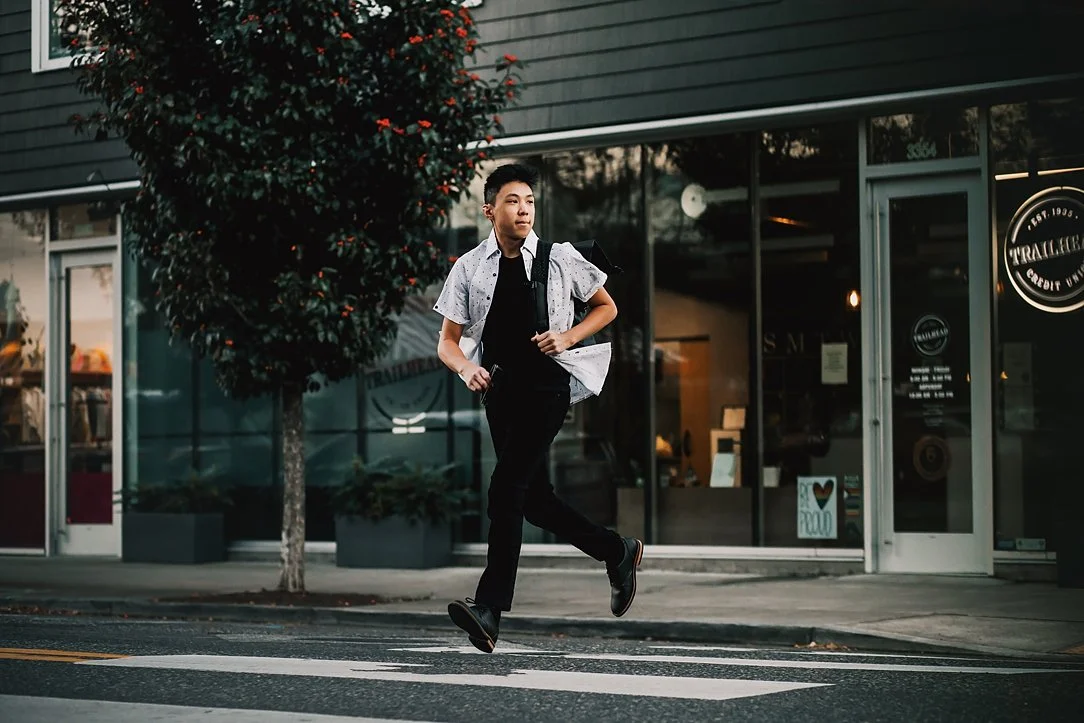 A teenage boy running across a crosswalk in front of modern storefronts during a senior portrait session in Portland.