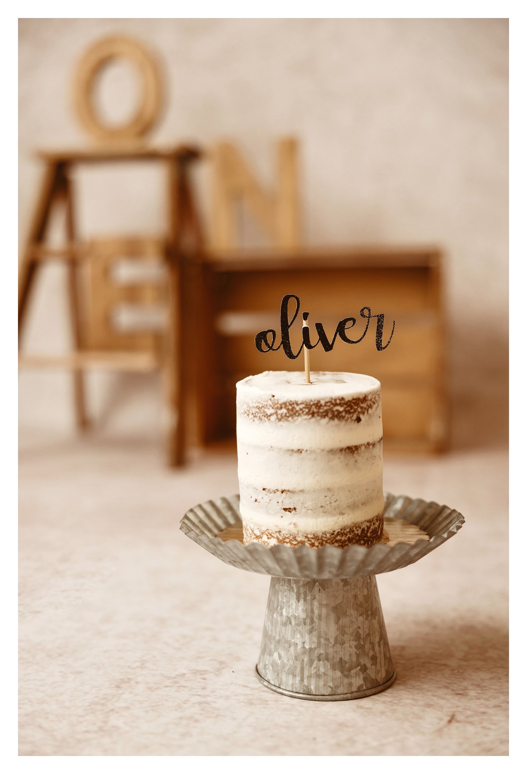 A simple cream layered naked cake on a galvanized metal stand with a black script topper reading Oliver, set against a blurred rustic wood backdrop.