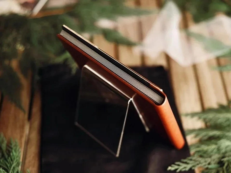 A side profile view of a thick, professional lay-flat photography album with a cognac-colored leather cover, displayed on a clear acrylic stand amidst greenery.