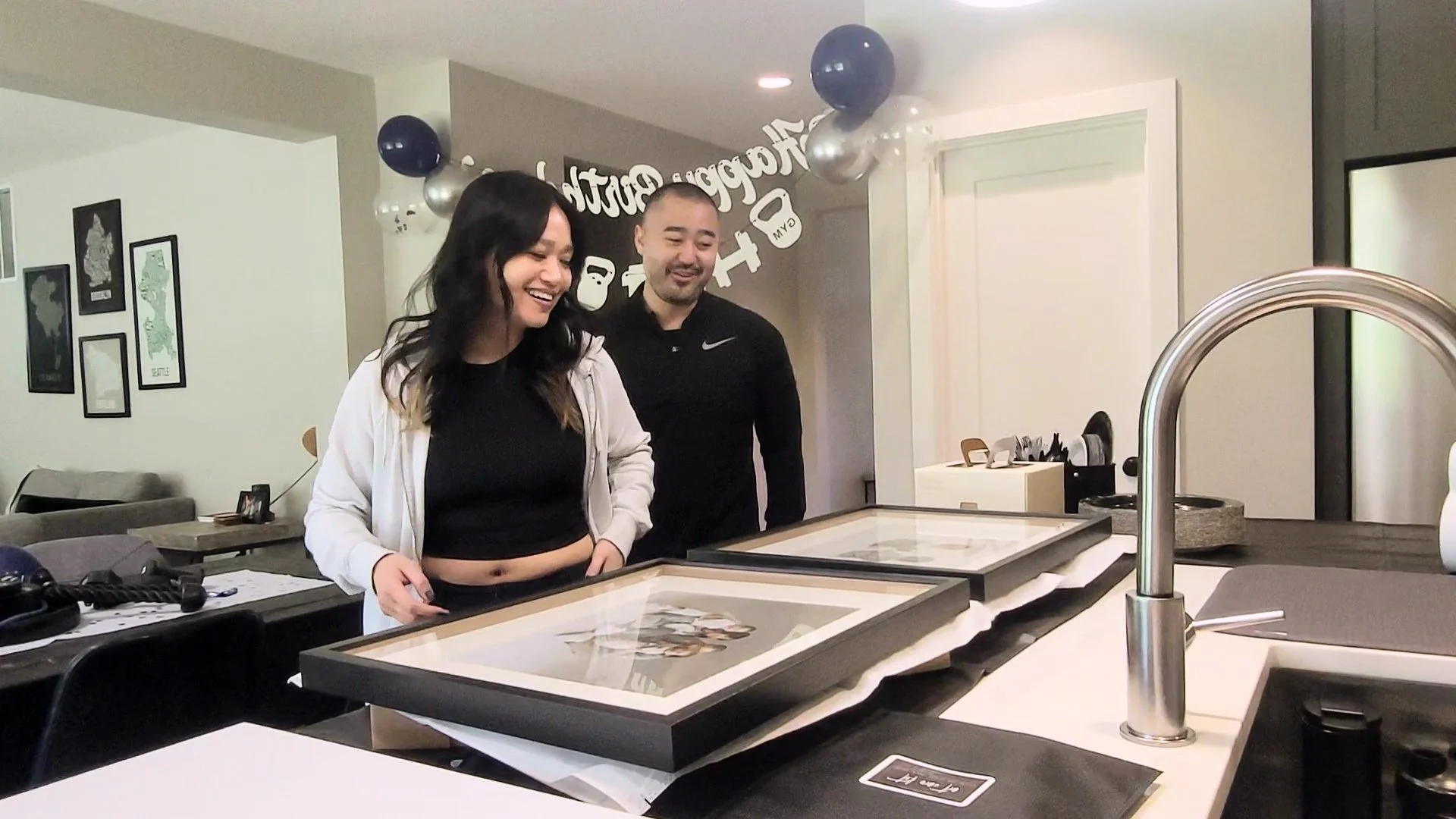 A happy young couple stands in their kitchen, smiling as they look down at two large, professionally framed family portraits. The scene highlights the excitement of a home gallery reveal and the high-quality finished products provided by a Portland f