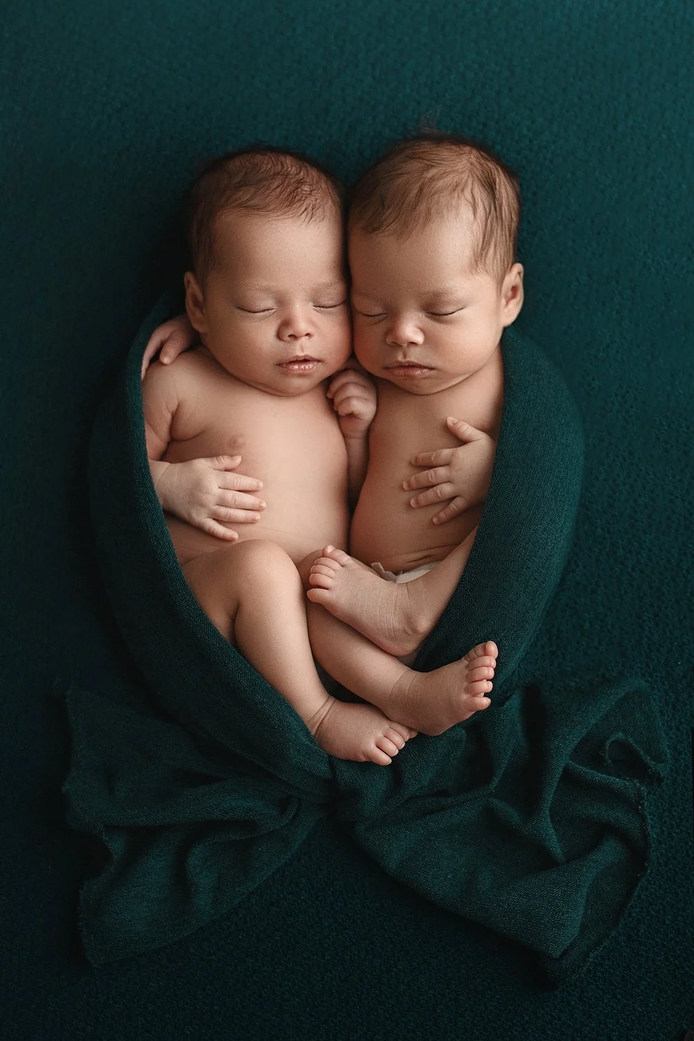 Fine-art studio portrait of twin newborn babies swaddled together in emerald green, sleeping against a matching textured backdrop in West Linn, Oregon.
