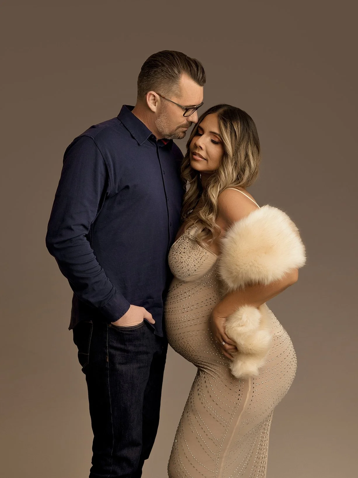 Sunnymelon Photography ultra-glamorous couples maternity portrait. An expectant mother in a form-fitting rhinestone-embellished gown and faux fur shawl poses with her partner, gently cradling her pregnant belly in a luxurious studio setting.
