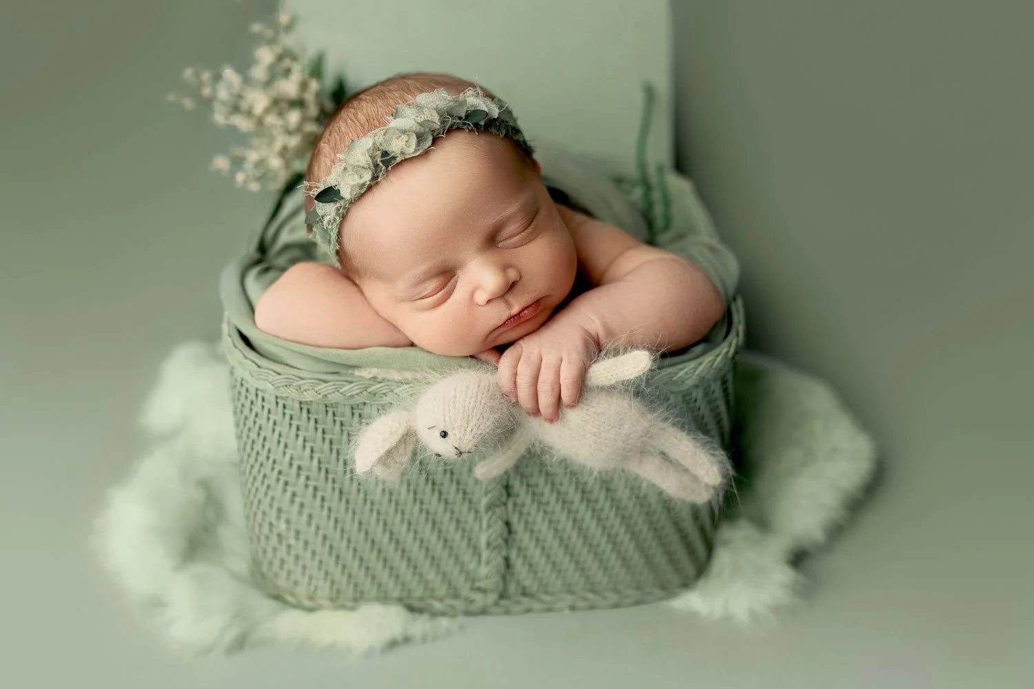 A serene fine-art studio portrait of a sleeping newborn baby nestled inside a sage green woven basket in West Linn, Oregon. Wearing a floral headband and holding a tiny knit bunny, the baby is captured in a soft, monochromatic setup.