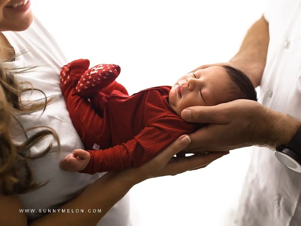 Close-up of a sleeping newborn baby in a red onesie being cradled by the hands of both parents in a bright studio.