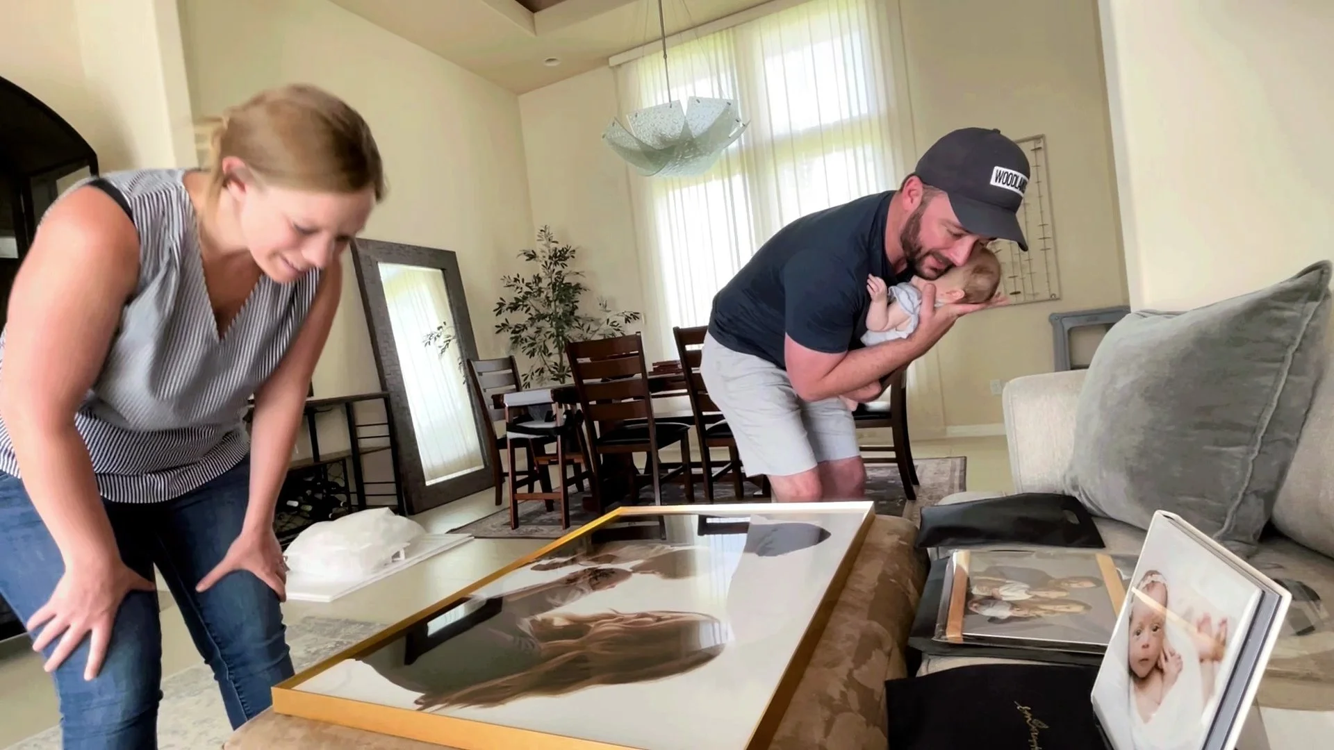 A candid, heartwarming scene in a bright living room where a mother and father are viewing newly printed professional photographs of their baby. The father, wearing a baseball cap, affectionately holds the infant while looking down at a large, gold-f