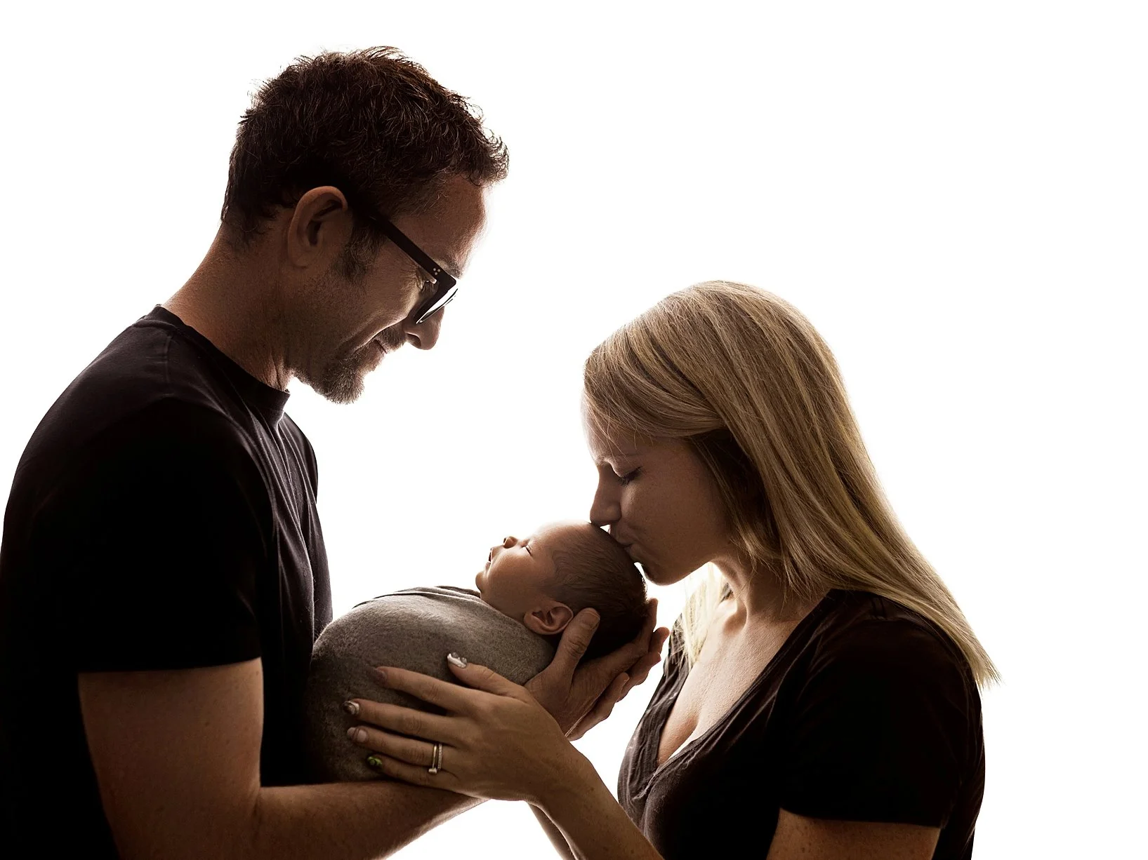 A tender high-contrast family photograph featuring a father on the left and a mother on the right, both in dark shirts, cradling their newborn infant together. The baby is peacefully asleep and swaddled in a soft grey wrap. The mother leans forward t