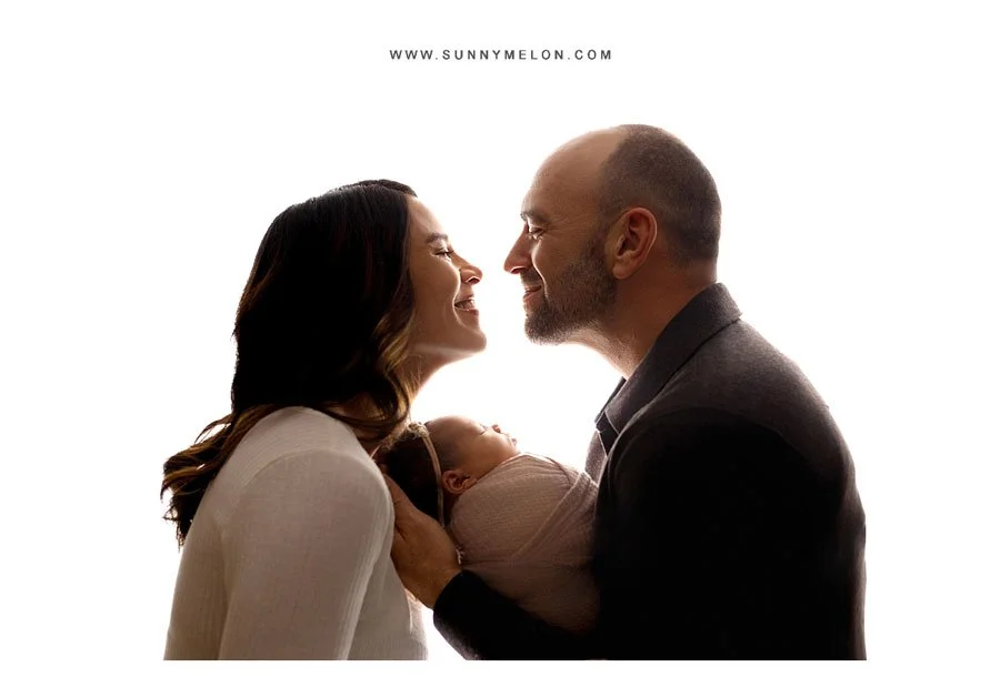 A back-lit studio portrait of a mother and father facing each other and smiling, while the father cradles their swaddled newborn baby against a bright white background.
