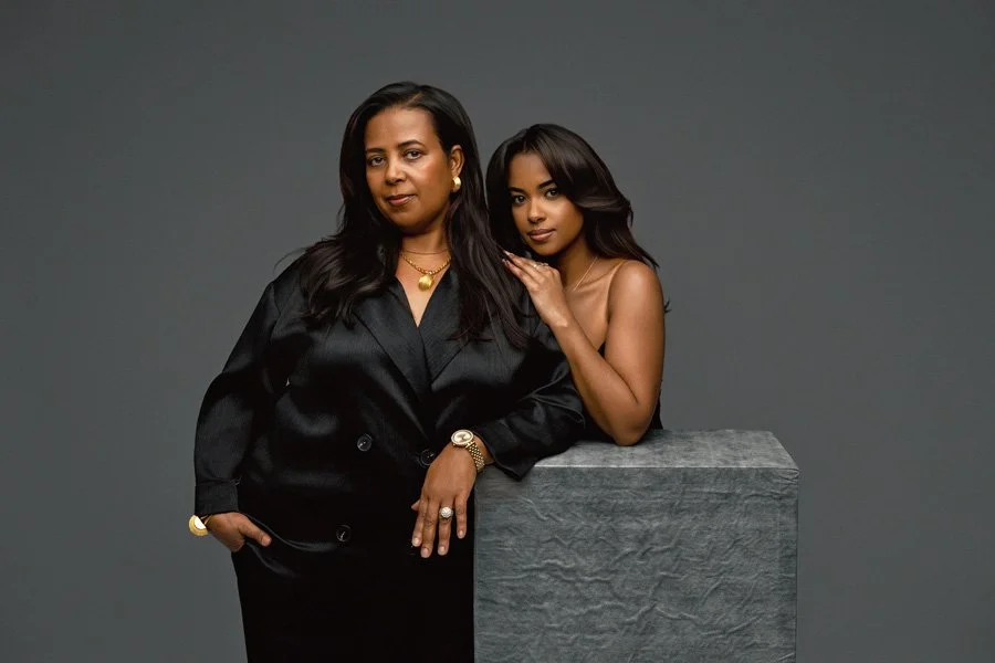 A high-end studio portrait of a mother in a black silk blazer and her daughter leaning on her shoulder, both posed against a solid grey background with a stone-textured pedestal.