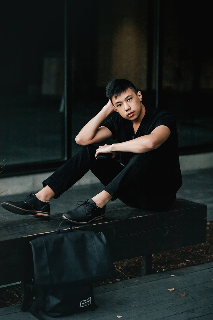 A high school senior boy in an all-black outfit posing on a dark bench for an urban lifestyle portrait session in downtown Portland.