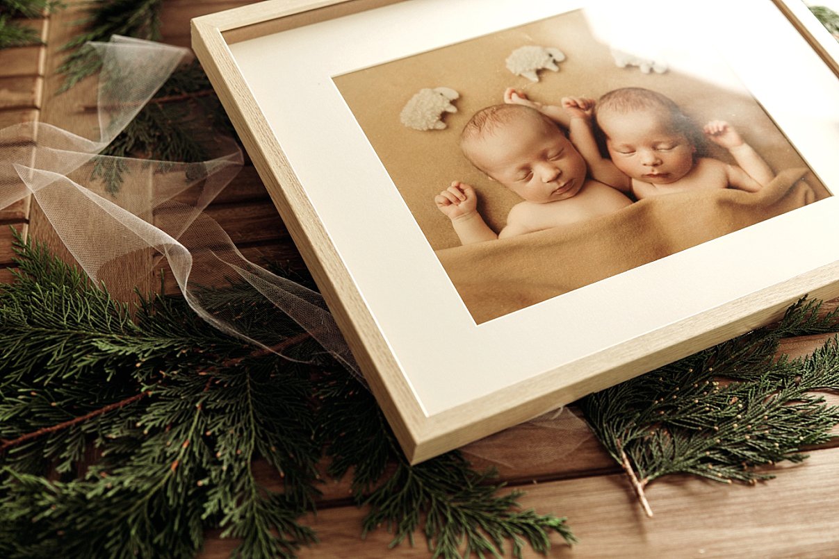 A high-quality framed fine art print of twin newborn babies sleeping under a tan blanket, displayed on a wooden surface with evergreen branches and delicate white tulle.