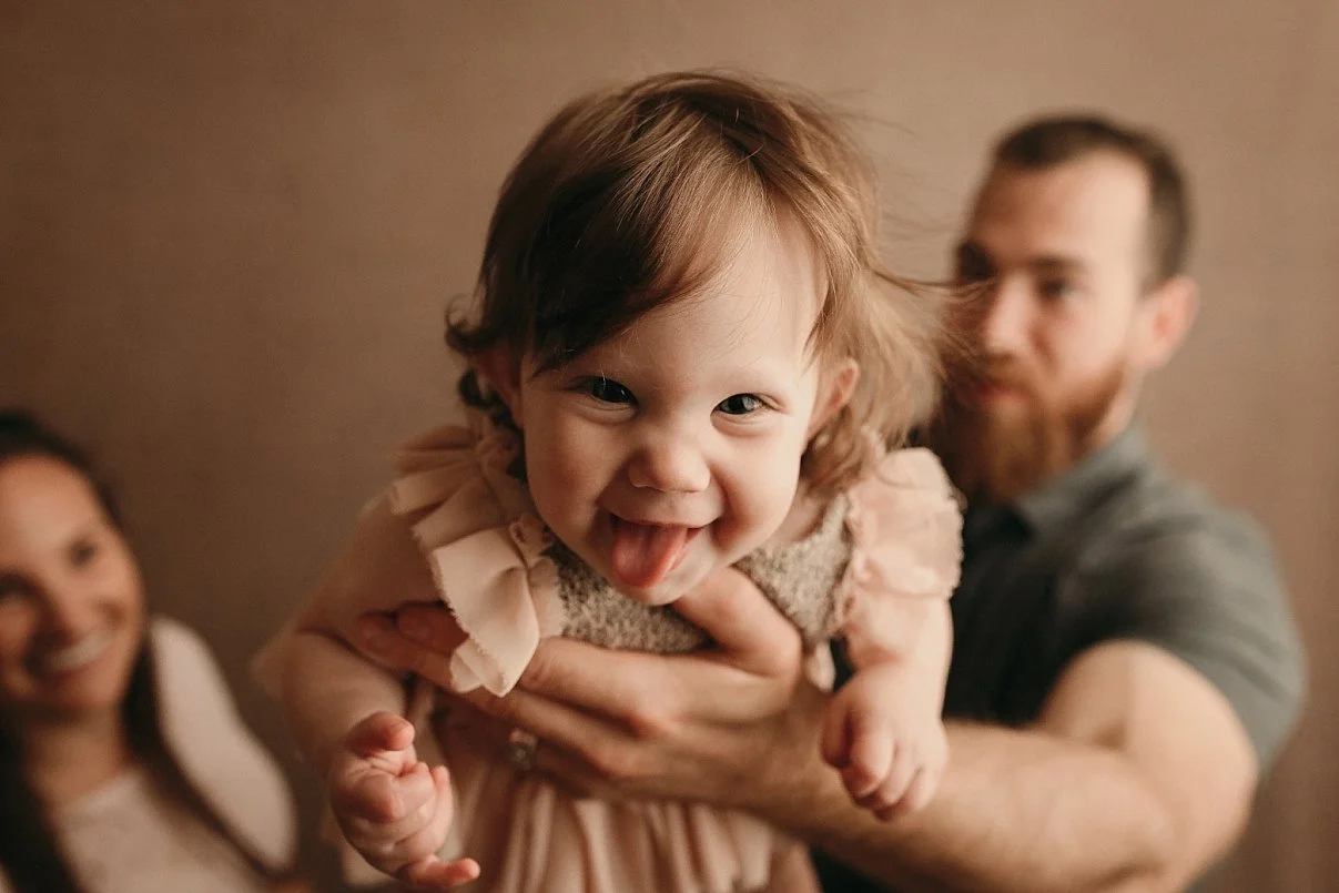 Joyful baby sticking out tongue, held by dad with mom in the background, capturing a happy family moment during a milestone sitter session in Portland, OR.