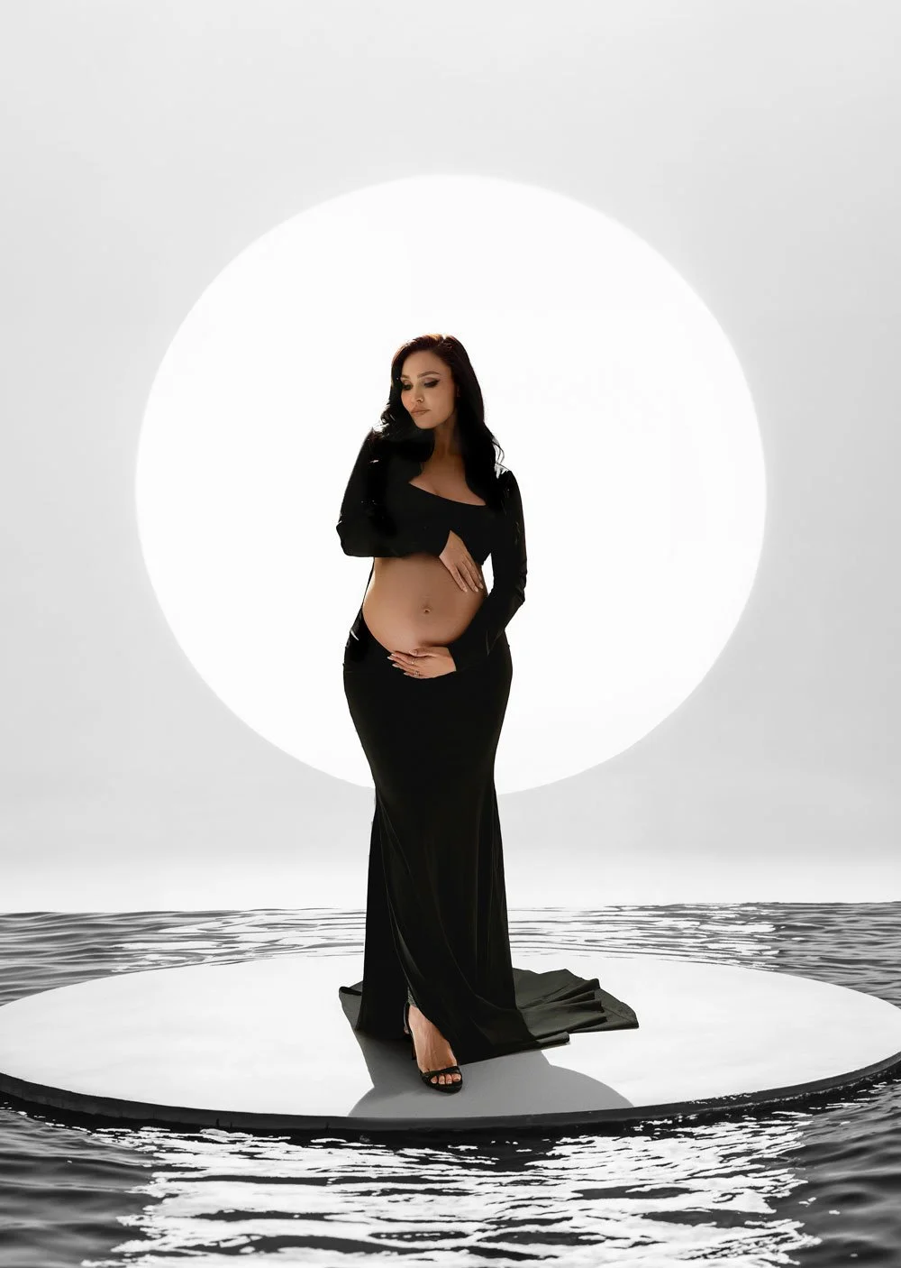 Fine-art studio maternity portrait of a pregnant woman in a black skirt and crop top on a circular platform over water with a large backlit halo backdrop in West Linn, Oregon.