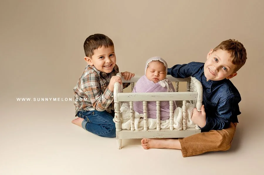 Two young boys smiling and posing next to their newborn sibling who is swaddled in purple and sleeping in a vintage-style miniature white crib, set against a warm cream background.
