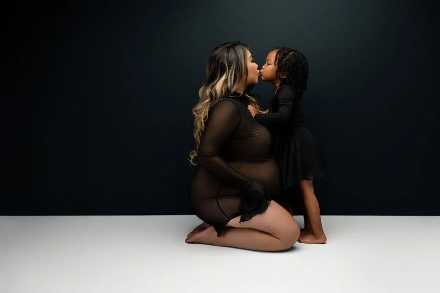 A professional studio maternity portrait of a pregnant woman in a sheer black outfit kneeling and sharing a kiss with her young daughter against a dark, minimalist background.