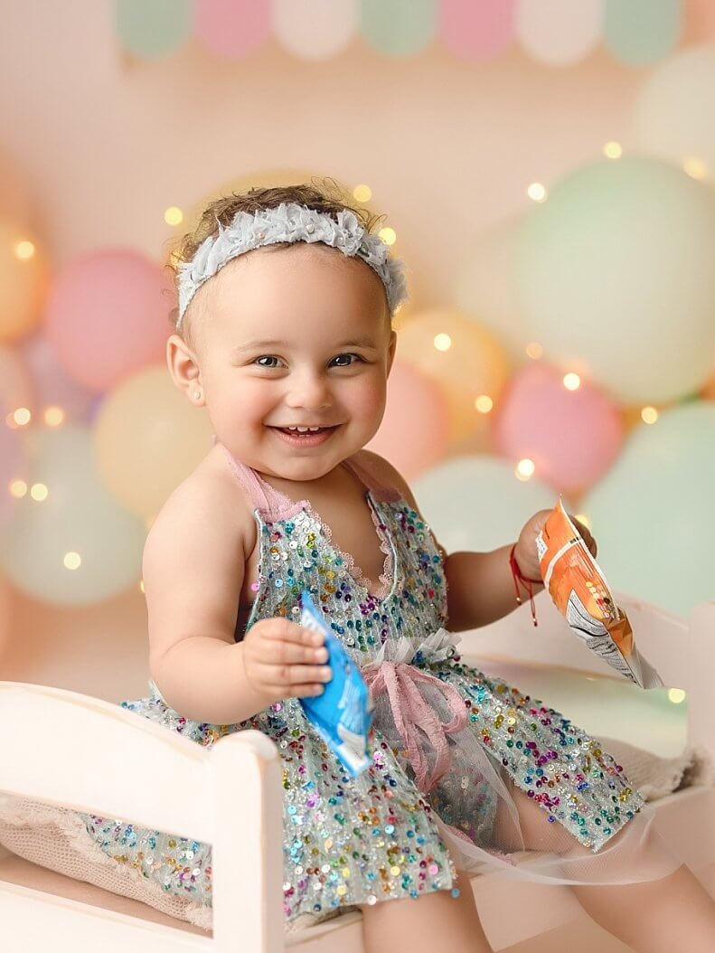 A smiling baby girl in a colorful sequin dress and grey floral headband sitting on a white bench during a first birthday photoshoot with a pastel balloon backdrop.