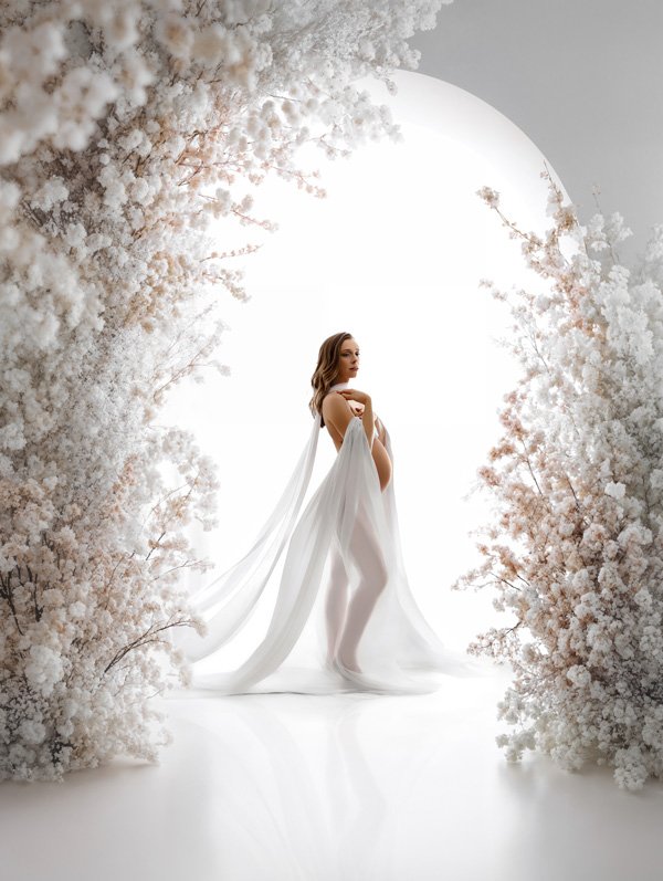 A pregnant woman with wavy brown hair wearing a sheer white draped gown, standing in a profile pose within a large archway made of white flowers against a bright white light.
