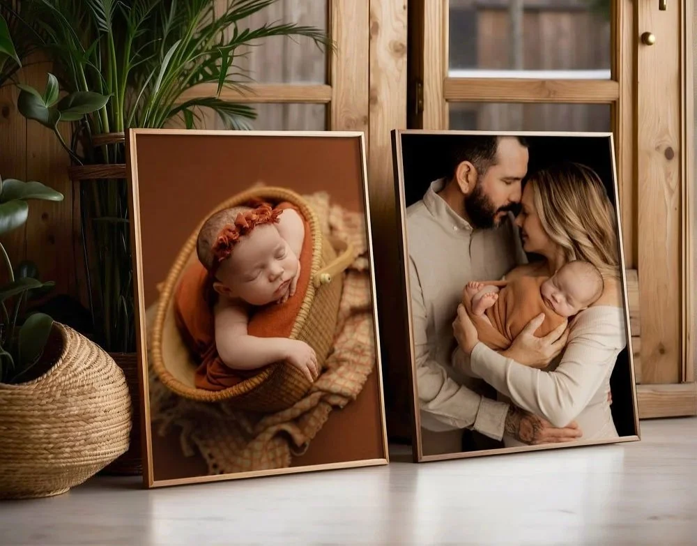 Two large, copper-toned metal frames containing professional fine-art portraits: one of a sleeping newborn in a basket and one of parents holding their baby, leaning against a wooden wall next to a potted plant.
