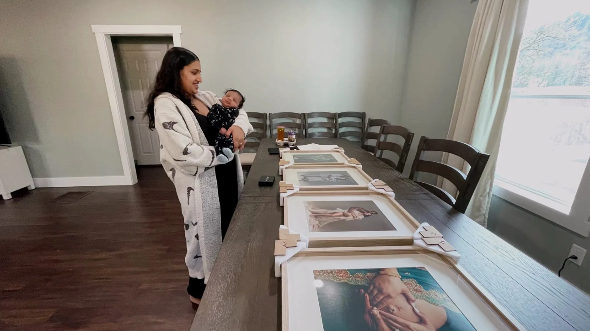 A woman holding a sleeping newborn baby while standing next to a dining table lined with large, framed maternity portraits.