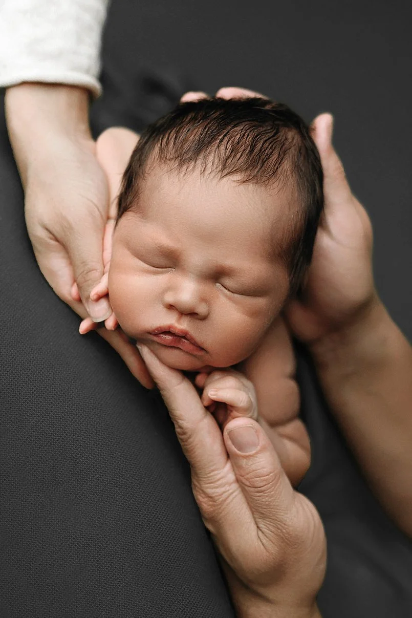 A tender studio photograph capturing a sleeping infant with dark hair. The baby's head is framed and supported by the hands of two adults, highlighting the scale and fragility of the newborn. The lighting is soft and directional, emphasizing the baby