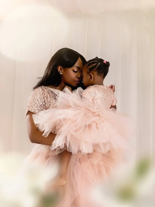 A mother holding her young daughter close, both wearing matching rose gold sequin tops and voluminous blush pink tulle skirts in a soft, ethereal studio setting.