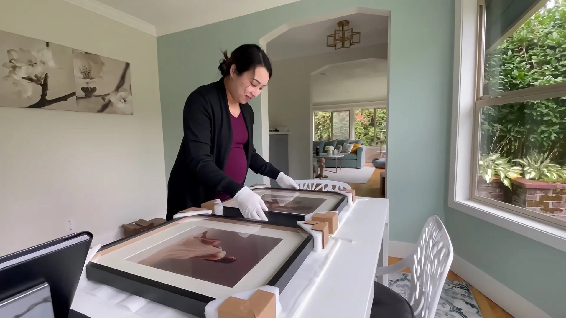 A client wearing white gloves carefully unboxing large, custom-framed maternity portraits on a white table in a bright, modern home studio.