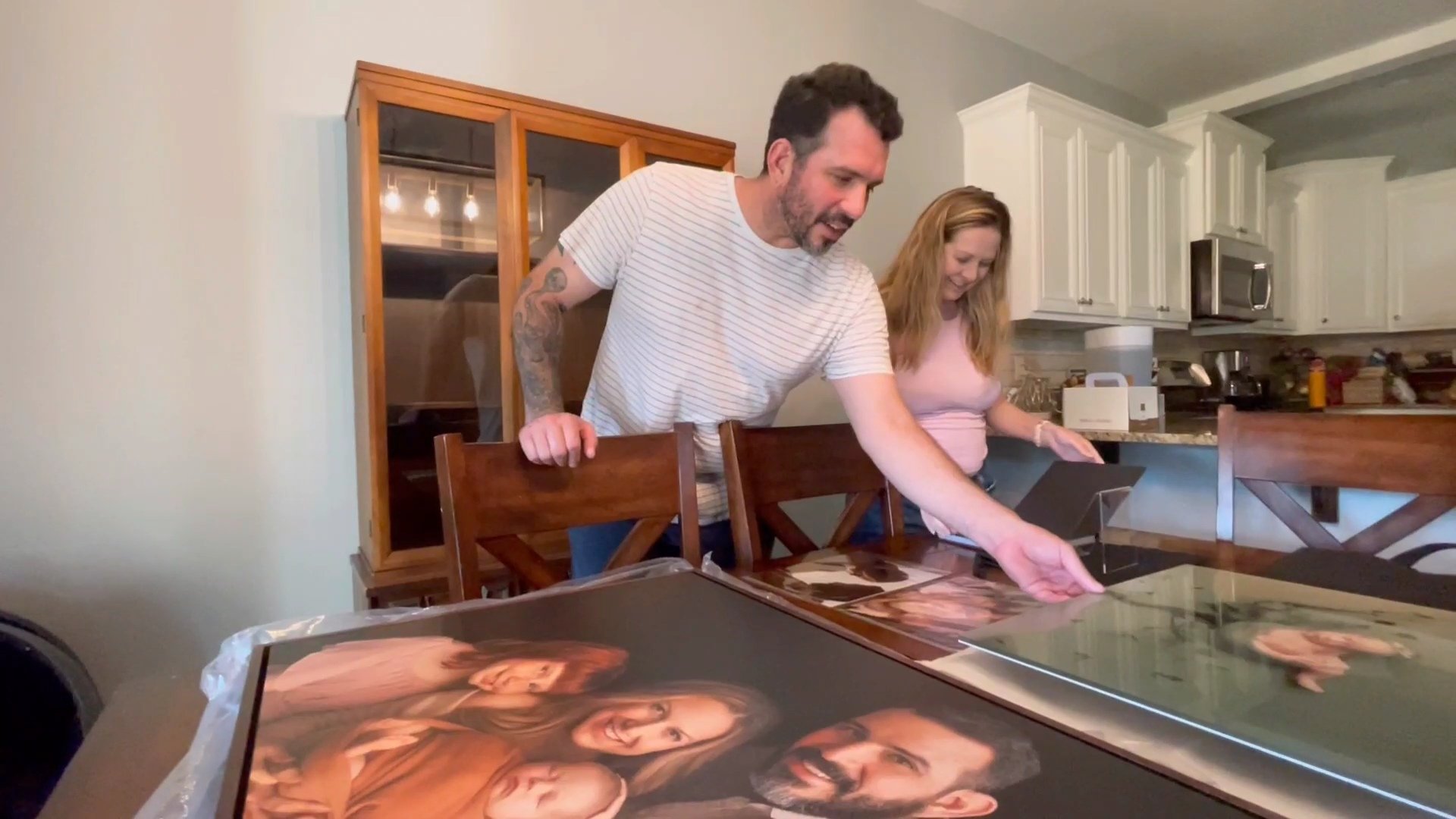 A man and woman standing in a dining area, excitedly looking at a large, framed family portrait and several smaller photography prints laid out on a wooden table.