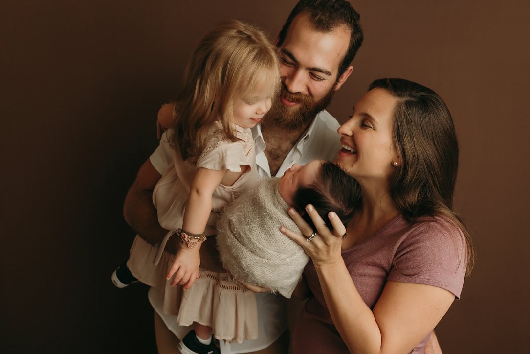 A professional newborn family portrait by SunnyMelon Photography featuring a mother, father, and toddler daughter admiring a newborn baby. The baby is snugly wrapped in a neutral, fuzzy mohair swaddle and held by the mother. The family is dressed in 