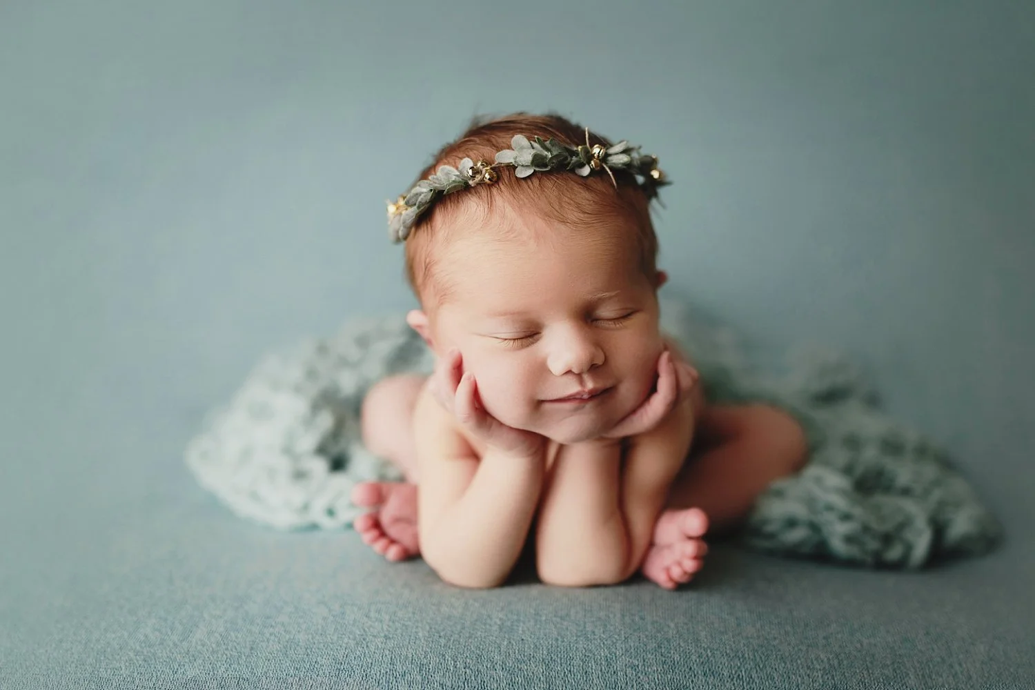 A tranquil newborn photography shot featuring an infant peacefully sleeping. The baby is positioned in a classic 'froggie' pose, with their head gently resting on their small hands. A dainty headband made of small green leaves and tiny white berries 