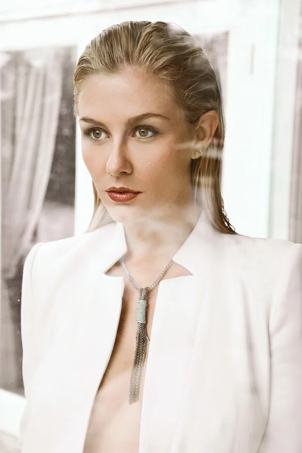 Close-up editorial portrait of a woman with slicked-back hair wearing a white architectural blazer and a silver tassel necklace, shot through a reflective glass pane.