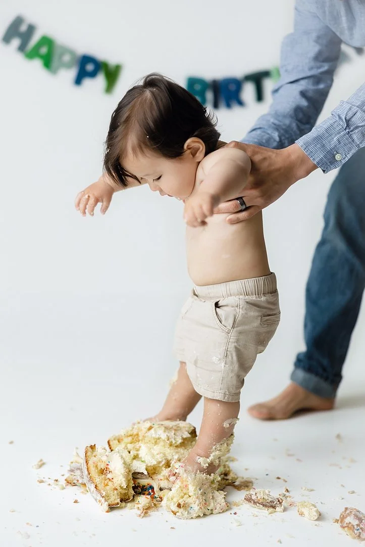This dynamic first birthday action shot captures a unique and candid moment during a professional cake smash session. The toddler, wearing neutral tan shorts, is being supported by an adult as he playfully plants his feet into the remains of a large 