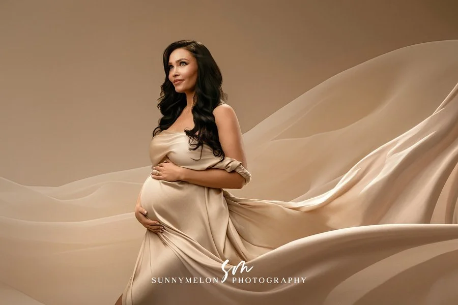 A pregnant woman with long dark hair posing in a champagne-colored satin drape against a matching neutral fabric backdrop in a professional photography studio.