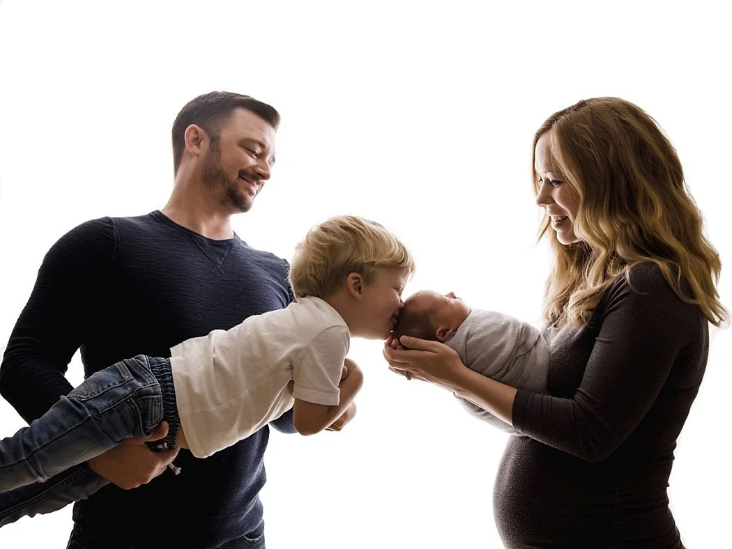 A backlit studio family portrait against a bright white background, featuring a father holding his young son who leans forward to kiss a swaddled newborn baby held by the mother.