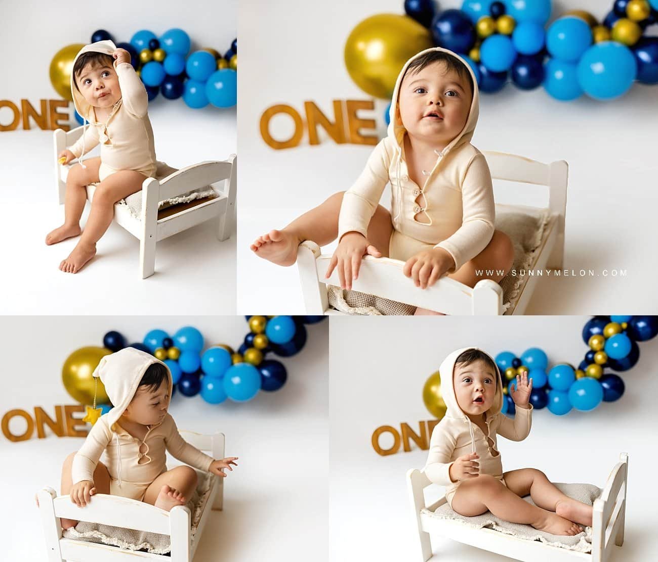 Toddler boy celebrating first birthday with blue and gold balloon arch and wooden "ONE" sign in a photography studio.