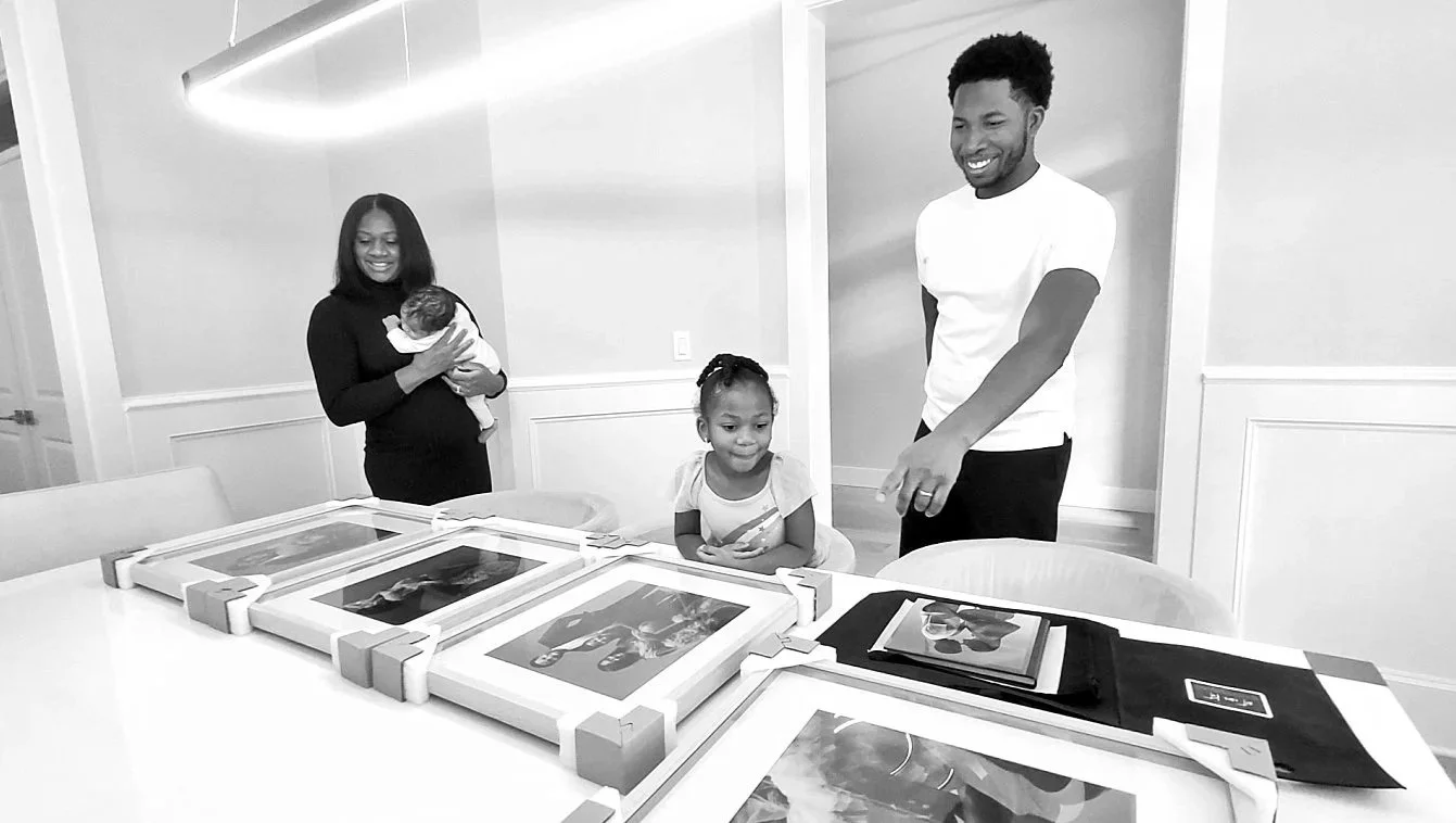 A family, including a mother holding a baby, a father, and a young daughter, smiling at several large, matted, and framed portraits laid out on a white dining table.