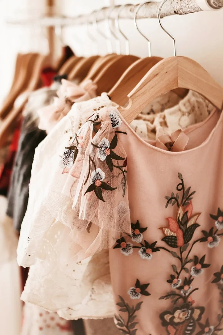 This detail shot showcases a boutique client closet, featuring a curated selection of delicate, high-end children's clothing for studio sessions. The focus is on a stunning pink dress with intricate floral embroidery and soft tulle sleeves, hanging n