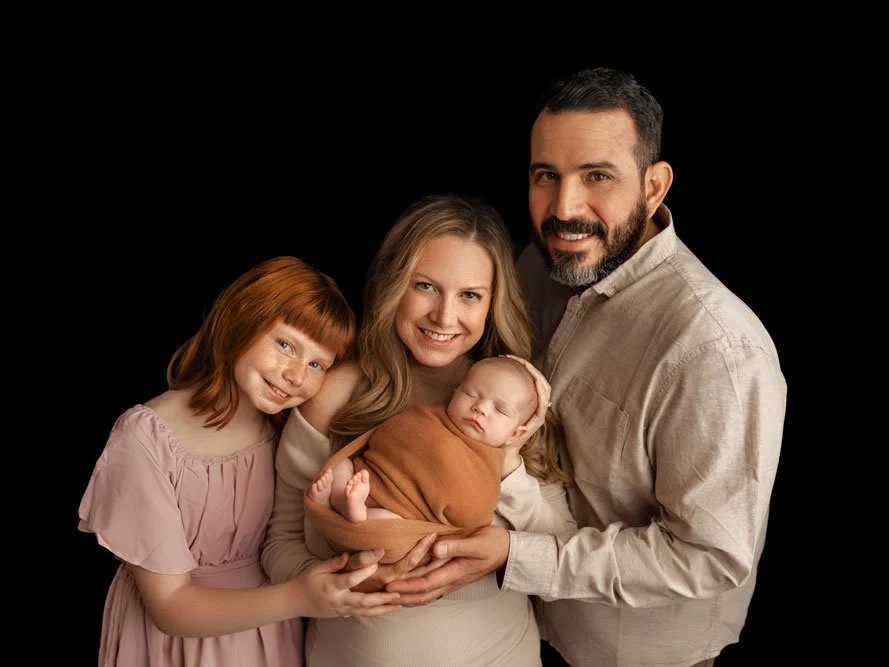 A professional studio newborn family portrait featuring a mother, father, and young daughter posing against a solid black background. The mother holds a sleeping newborn baby wrapped in a warm terracotta swaddle, while the father and older sister lea