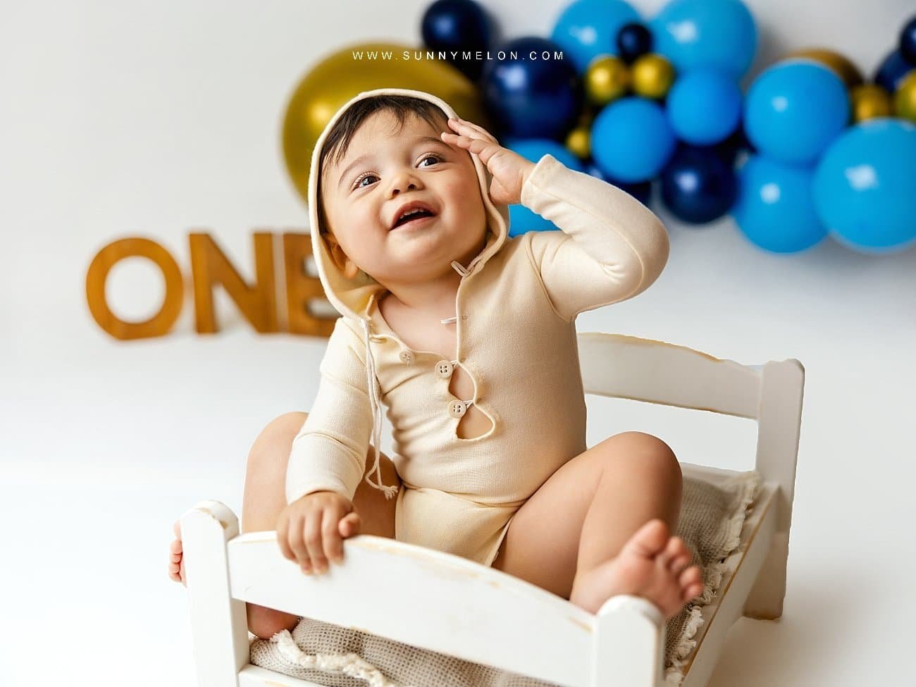 Smiling toddler boy in a cream hooded romper sitting on a white prop bed with blue and gold balloons.